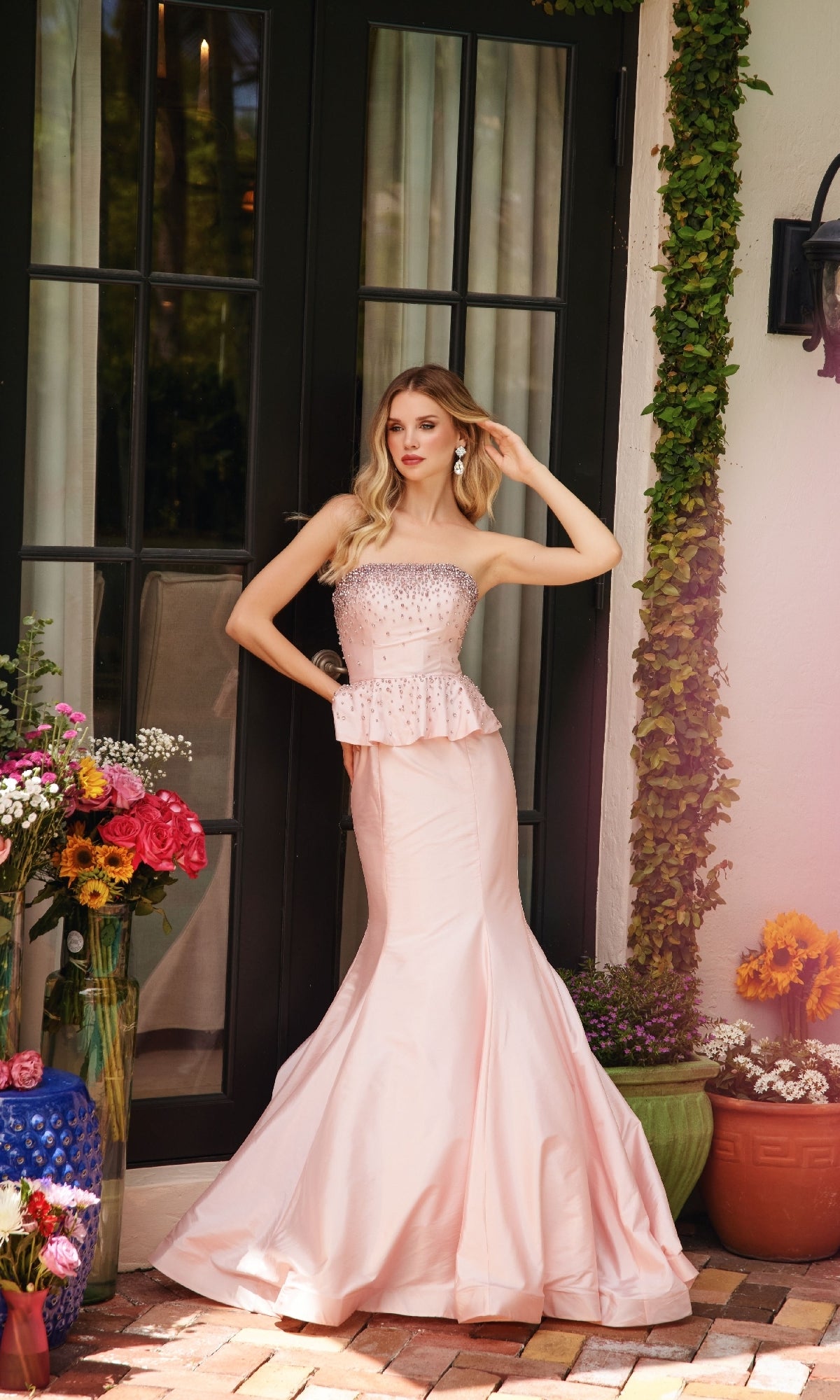 A woman in the Ava Presley 40092 Strapless Peplum Prom Dress stands outside by glass doors, surrounded by potted plants and colorful flowers.